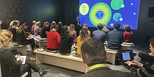 Referent steht vor Publikum und zeigt auf einen Bildschirm